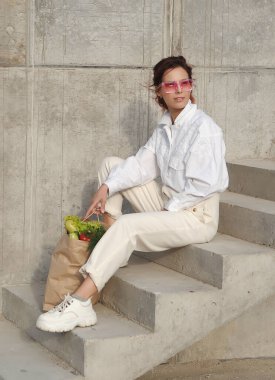 Tarz sahibi güzel bir kadın, açık havada poz veren sebzelerle dolu bir kağıt çanta tutuyor..