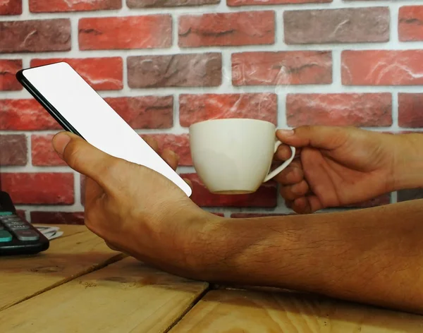 White screen smartphone in the hands of a young businessman who is dealing in a coffee shop behind a brick wall