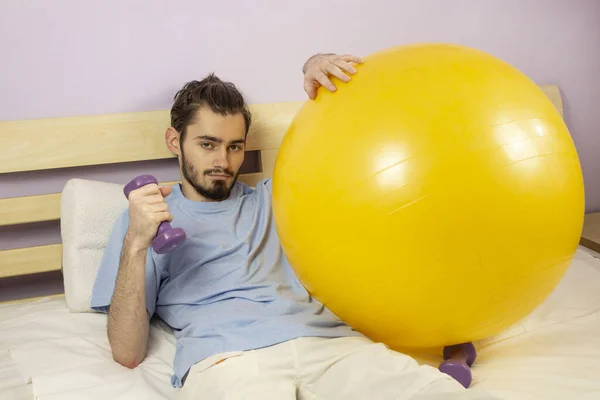 Sport at home. Go in for sports at home during quarantine. A young guy holds a dumbbell in bed, next to a fitball. To stay home.