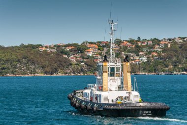 Sydney, Avustralya - 11 Aralık 2009: Karoo Kemble Limanı 'nın siyah-beyaz römorkörü derin mavi körfez sularında, ufukta kıyı çizgisi açık mavi gökyüzü altında kırmızı çatılı evlerle dolu..