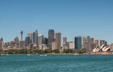 Sydney, Australia - December 11, 2009: Opera House as small building with wide cityscape of office towers under blue sky and azure bay water in front.