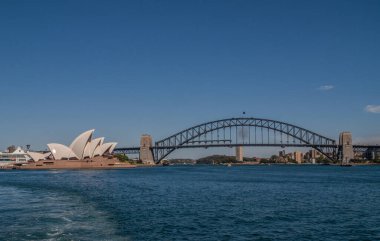 Sydney, Australia - December 11, 2009: Opera House with full black Harbour bridge and its anchor towers over deep blue bay water and under lighter blue sky. 