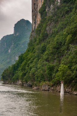 Guilin, China - May 10, 2010: Along Li River. White and red navigation beacon standing in greenish water at foot of brown vertical cliff, partly covered by green foliage. Other mountains and rainy sky.