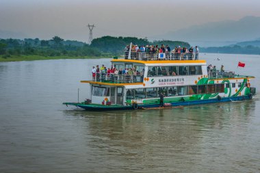 Guilin, China - May 10, 2010: Along Li River. White-orange-green overloaded tourist sightseeing boat on brown water with green shoreline under brown foggy sky. Vendor on raft.