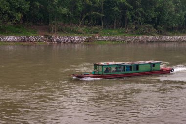 Guilin, China - May 10, 2010: Along Li River. Green-brown transport boat on dark brown water. Forest covered shoreline with reinforced edge and stairway.