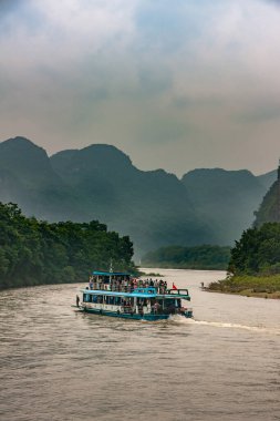 Guilin, China - May 10, 2010: Along Li River. White-blue overloaded tourist sightseeing boat meanders with brown water Green shoreline and karst mountains under brown foggy sky.