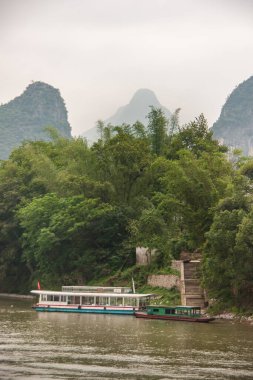 Guilin, China - May 10, 2010: Along Li River. Ferry and transport vessel on brown water docked at stairs surrounded by green foliage with karst mountain tops in foggy gray sky as backdrop.