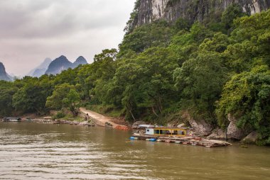 Guilin, China - May 10, 2010: Along Li River. Long Barrel supported raft with construction tools anchored near point where road reaches water. Green foliage shoreline. Karst mountains under silver sky.