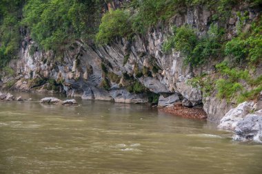 Guilin, China - May 10, 2010: Along Li River. Green foliage covers partly opening to cave in white-gray cliff on level with greenish water.