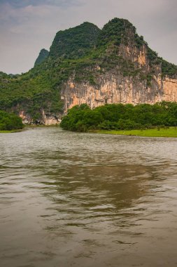 Guilin, China - May 10, 2010: Along Li River. Landscape with green forested karst mountains with brown-black vetical cliff under dark gray sky. Some trees, and Brown water up front.