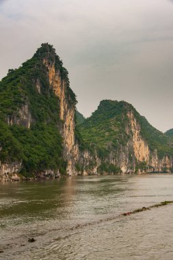 Guilin, Çin - 10 Mayıs 2010: Li Nehri boyunca. Gri gökyüzünün altında kahverengi-siyah vetik uçurumları olan yeşil ormanlı karst dağları. Önde yeşilimsi kahverengi su.
