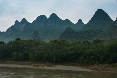 Guilin, Çin - 10 Mayıs 2010: Li Nehri boyunca. Mavimsi bulutların altında yeşil orman karst dağının manzarası. Kahverengi su ve ağaçlarla dolu kıyı şeridi önde..