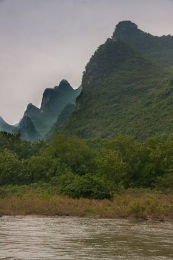 Guilin, Çin - 10 Mayıs 2010: Li Nehri boyunca. Gümüş bulutlar altında yeşil ormanlık karst dağının manzarası. Kahverengi su ve çalılar önde..
