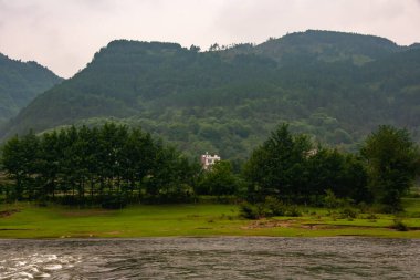 Guilin, Çin - 10 Mayıs 2010: Li Nehri boyunca. Karst Dağları 'nın önündeki, yağmurlu gökyüzünün altındaki gizli yüksek binaların manzarası. Ağaçlı yeşil kıyı şeridi. Koyu kahverengi su.