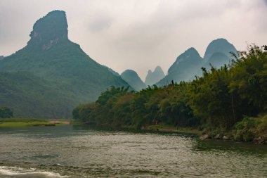 Guilin, Çin - 10 Mayıs 2010: Li Nehri boyunca. Özel şekilli karst dağ zirvesinin manzarası yağmurlu gökyüzünün altında. Ağaçlı yeşil kıyı şeridi. Koyu kahverengi su.