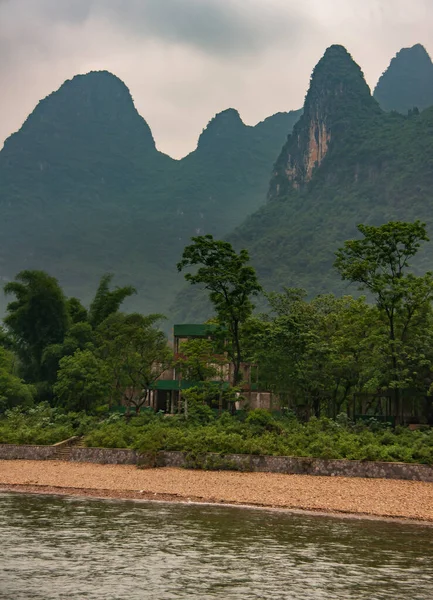 Guilin, China - May 10, 2010: Along Li River. Forested karst mountain range with rounded tops under gray clooudscape. Cubist mansion under construction hidden between trees on brown shoreline.