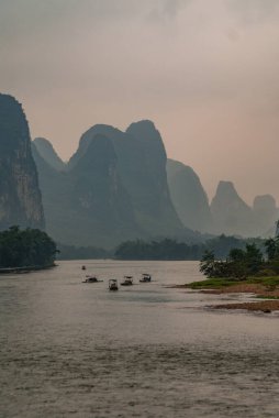 Guilin, Çin - 10 Mayıs 2010: Li Nehri boyunca. Karst sıradağları kahverengi bulutların altında su üzerinde 5 küçük teknenin yaklaştığı manzara. Sahilde yeşil ağaçlar.