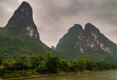 Guilin, Çin - 10 Mayıs 2010: Li Nehri boyunca. Kahverengi bulutların altındaki kıyılarda yüksek karst sıradağları olan bir manzara. Yeşil ağaçların arasından toprak yolun geçtiği kıyıda küçük tekneler bekliyor..