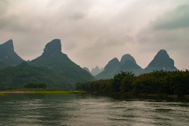 Guilin, Çin - 10 Mayıs 2010: Li Nehri boyunca. Koyu suyun arkasındaki gri bulutlar altında, daha açık yeşil kıyı şeridi ve yoğun ormanın altında birçok karst dağ zirvesi olan manzara..
