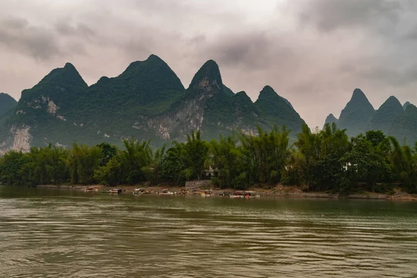 Guilin, China - May 10, 2010: Along Li River. Landscape with small ferry boats docked near steps between green trees in front of range of karst mountains under brown cloudscape.