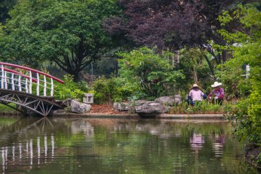 Guilin, China - May 11, 2010: Seven Star Park. 2 gardeners work behind pond and bow bridge in garden with their images mirrored in water.