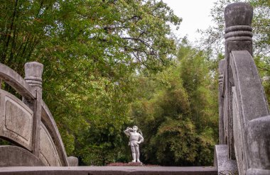 Guilin, China - May 11, 2010: Seven Star Park. White statue of boy in military uniform blowing the bugle in front of wall of green foliage as seen from over gray stone bow bridge.