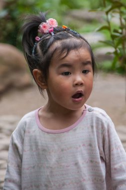 Guilin, China - May 11, 2010: Seven Star Park. closeup of little girl with wide open mouth is wondering. Fake flowers in her black hair. Light shirt and green foliage in back.