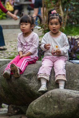 Guilin, China - May 11, 2010: Seven Star Park. Frontal closeup of 2 little girls sitting on dark gray boulder. Fake flowers in black hair. Light shirts and multicolor backdrop.