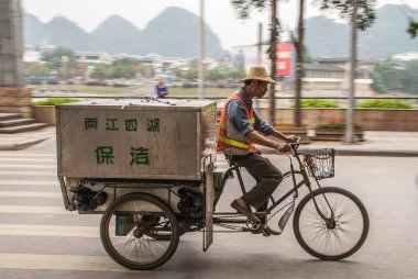 Guilin, Çin - 11 Mayıs 2010: şehir merkezi. Hasır şapkalı ve turuncu güvenlik yelekli bir adam arkada Karst Dağları ve Li Nehri boyunca büyük ulaşım araçlarıyla üç tekerlekli bisiklet sürüyor..