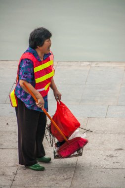 Guilin, Çin - 11 Mayıs 2010: şehir merkezi. Kırmızı-sarı güvenlik yeleği giyen kadın Li Nehri boyunca elinde temizlik aletleri ve kırmızı çantayla rıhtımı tarıyor..