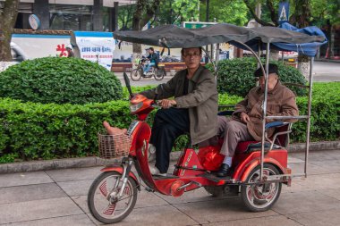 Guilin, Çin - 11 Mayıs 2010: şehir merkezi. Genç adam, yaşlı adamı kırmızı, siyah çatılı, üç tekerlekli scooter 'la, işlek caddenin kaldırımında taşıyor. Yeşil yaprak.