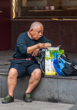 Şangay, Çin - 4 Mayıs 2010: Deniz mavisi giyinmiş yaşlı bir adam koyu gri kaldırımda oturur ve iş yerinin önündeki yeşil-beyaz karton üzerinde öğle yemeğini yer..