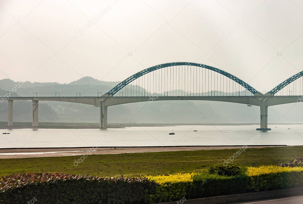 Chenjiawan, China - May 5, 2010: Bow bridge over Yangtze river under silver sky with green vegetation and yellow flowers on near shore but covered green hills in fog on other shore.