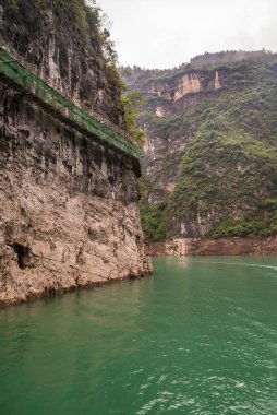 Wuchan, Çin - 7 Mayıs 2010: Daning Nehri üzerindeki Dawu veya Misty Gorge. Zümrüt yeşili suyun üzerinde kahverengi-siyah bir uçurum üzerine inşa edilmiş yeşil gözenekli çitli uzun tahta patika yolu.. 
