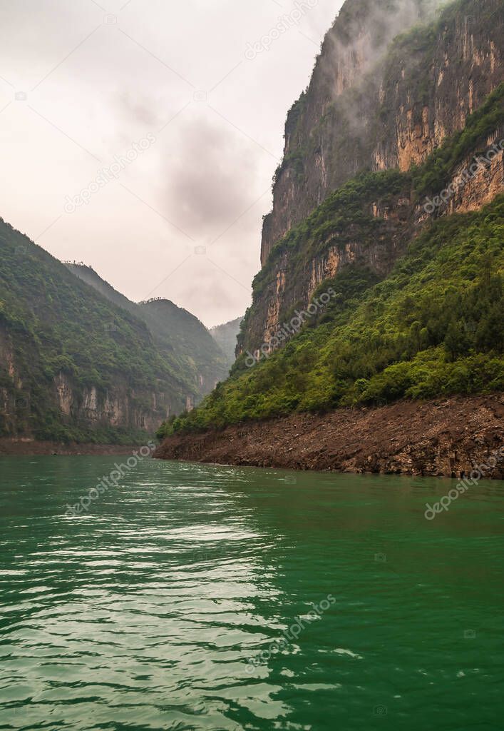 Wuchan, China - May 7, 2010: Dawu or Misty Gorge on Daning River. Portrait of Bend in canyon with brown rocky cliffs, green foliage on top, emerald green water, and misty cloudscape.