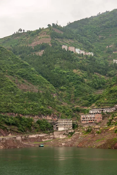 Shengli Street, China - May 6, 2010: Xiling gorge on Yangtze River. Rows of houses near green water shoreline and on top on  green tree covered mountain slope. Silver sky. One ferry boat.