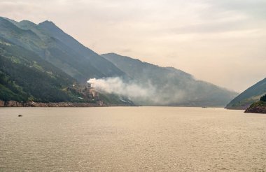 Xinling, Çin - 6 Mayıs 2010: Xiling vadisi Yangtze Nehri üzerinde. Duman, kıyı şeridi boyunca uzanan büyük çimento fabrikasından ve kahverengi gökyüzünün altındaki yeşil dağlardan kaçar. Geniş kahverengi su.