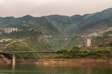 Xinling, Çin - 6 Mayıs 2010: Xiling vadisi Yangtze Nehri üzerinde. Yangtze 'ye giden 2 yol köprüsü. Arkasına serpilmiş binalar serpiştirilmiş yüksek dağlar. Kahverengi gökyüzü ve yeşil su.
