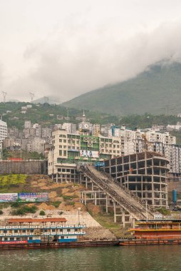 Xinling, Çin - 6 Mayıs 2010: Xiling vadisi Yangtze Nehri üzerinde. Sahil şeridindeki inşaat alanının arkasındaki saat kulesinde yeşil dağlar ve şehir manzarası üzerine kayıklar, reklam panoları ve bulutlar iniyor..