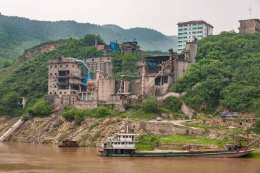 Huangqikou, Chongqing, Çin - 8 Mayıs 2010: Yangtze Nehri. Kıyı şeridindeki yeşil yapraklar arasında kahverengi suya demirlenmiş mavnayla kahverengi taş yıkıcı fabrika kurulmuş. Arkada büyük bir bina var..