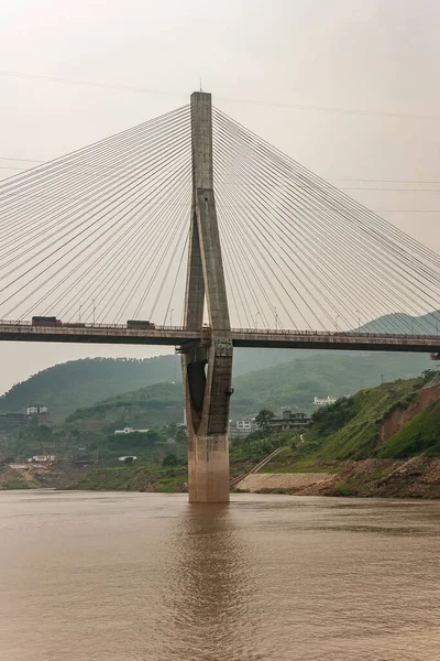 Huangqikou, Chongqing, China - May 8, 2010: Yangtze River. 1 of 2 diamond shaped pylons of G319 suspension bridge with trucks. Brown water and green hills with sprinkled white buildings in back.