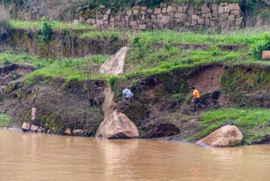 Fengdu, Chongqing, Çin - 8 Mayıs 2010: Yangtze Nehri. Siyah elbiseli çocuklar koyu toprak ve bej kayalar üzerinde oynuyorlar. Üzerinde yeşil bitki örtüsü ve önünde kahverengi su var..