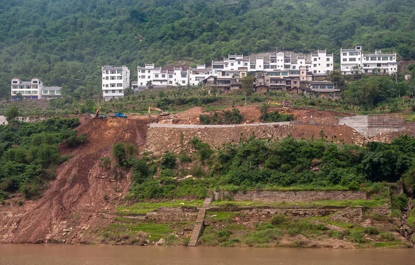 Fengdu, Chongqing, China - May 8, 2010: Yangtze River. White housing row near green forest on top of shoreline where colapsed road is being repaired with trucks and cranes present. Brown water.
