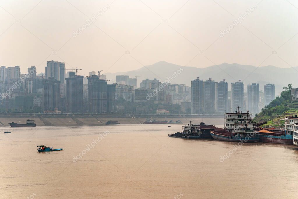 Fengdu, Chongqing, China - May 8, 2010: Yangtze River. Cityscape of high buildings on shoreline behind wide brown water with some vessels sailing and moored. Smoggy sky.