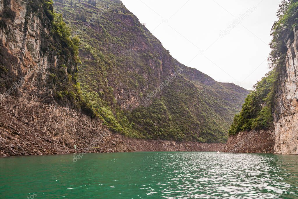 Wushan, Chongqing, China - May 7, 2010: Mini Three Gorges on Daning River. Landscape of wide canyon with high cliff brown rock mountains under silver sky and emerald green water up front.