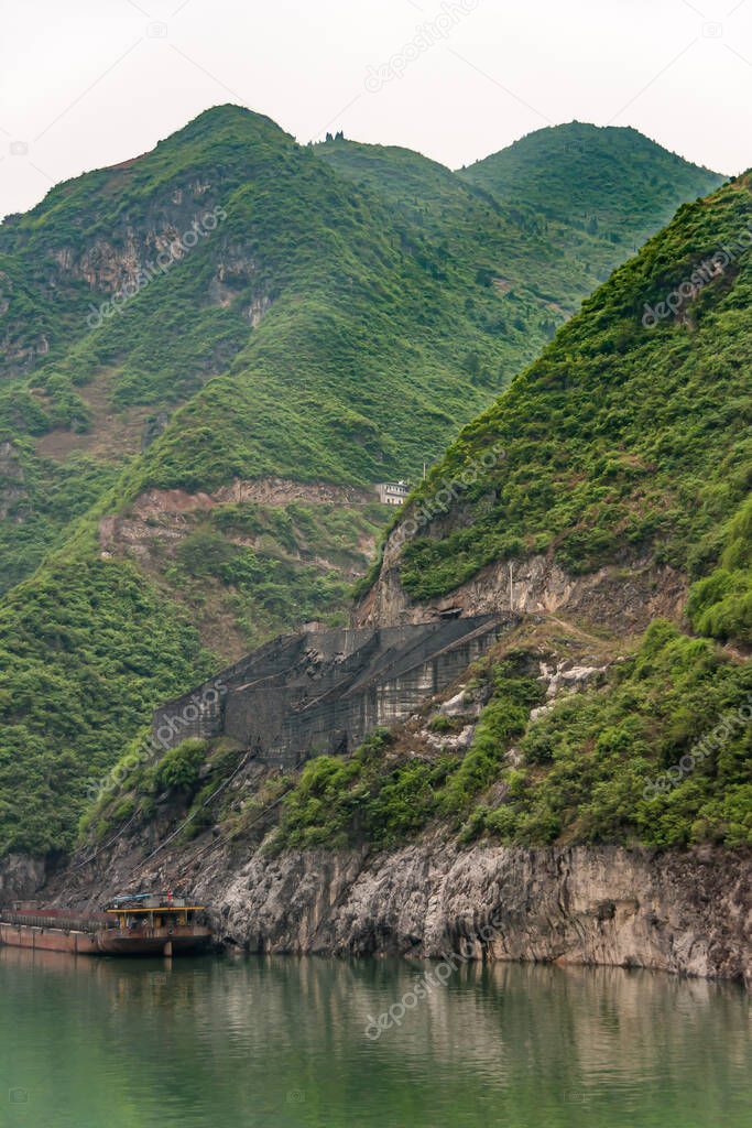 Guandukou, Hubei, China - May 7, 2010: Wu Gorge in Yangtze River. Wall structure above coal loading dock on shoreline of green foliage covered mountain  with barge present on green water.