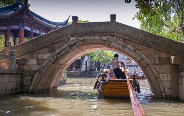 Tongli, Jlanangsu, Çin - 3 Mayıs 2010: Kanal manzarası. Kahverengi ahşap yüzme teknesinin kaptanı, kahverengi-yeşil suların altında mavi çatı kiosku yakınlarındaki gri taş köprünün altında yelken açarken başını eğmeli..