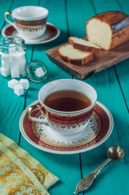 Kesme tahtasının üzerinde dilimlenmiş bir pasta, şeker küpleri ve iki porselen çay bardağı olan Tiffany renginde ahşap bir masa.