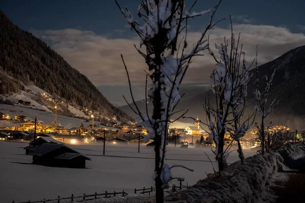 Paisagem noturna de inverno com vista para a cidade tirolesa austríaca de Neustift e a igreja de Pfarre no pano de fundo de montanhas e nuvens. Noite gelada com nevoeiro no vale — Fotografia de Stock