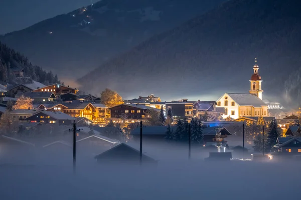 Paisagem noturna de inverno com vista para a cidade tirolesa austríaca de Neustift e a igreja de Pfarre no pano de fundo de montanhas e nuvens. Noite gelada com nevoeiro no vale — Fotografia de Stock